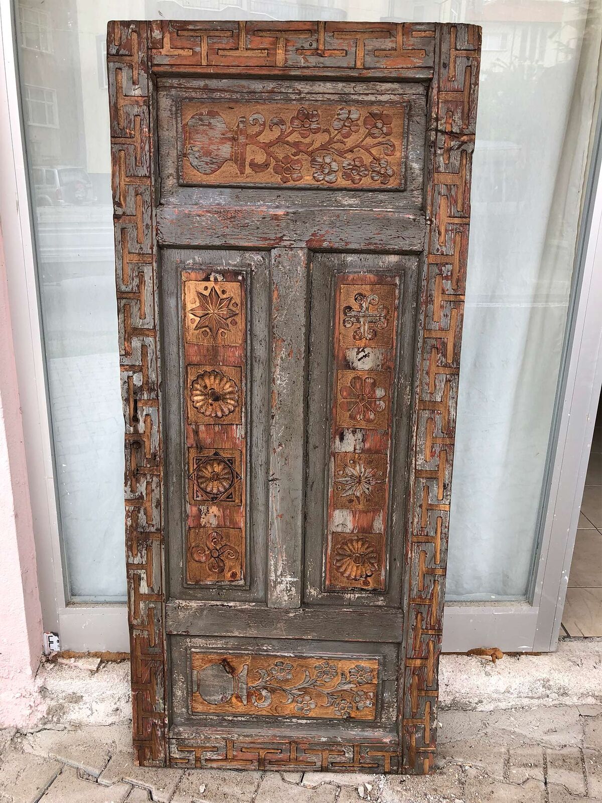 Carved Door with Inset Panels — tall brown wooden door with carved inset panels leaning against a shop window